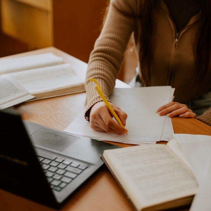 Eine Person schreibt mit einem Bleistift auf Papier neben einem Laptop und Büchern.